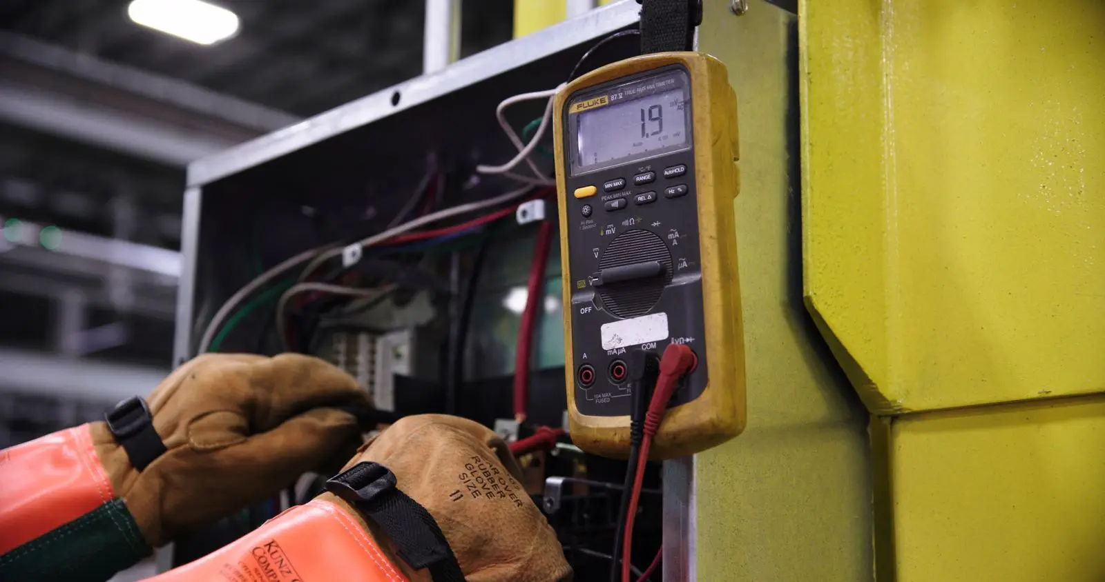 Gloved hand holding a digital clamp meter up to electrical equipment