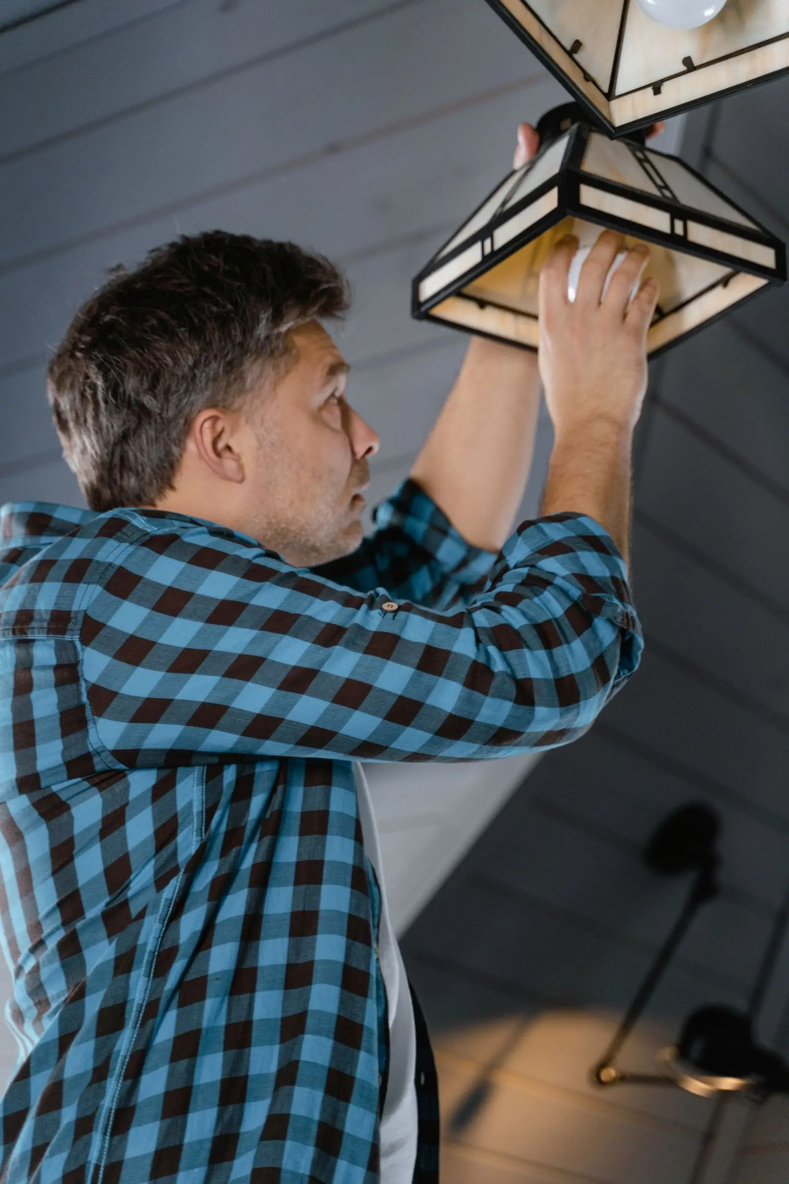 Man in a plaid shirt holding a light fixture up to a ceiling