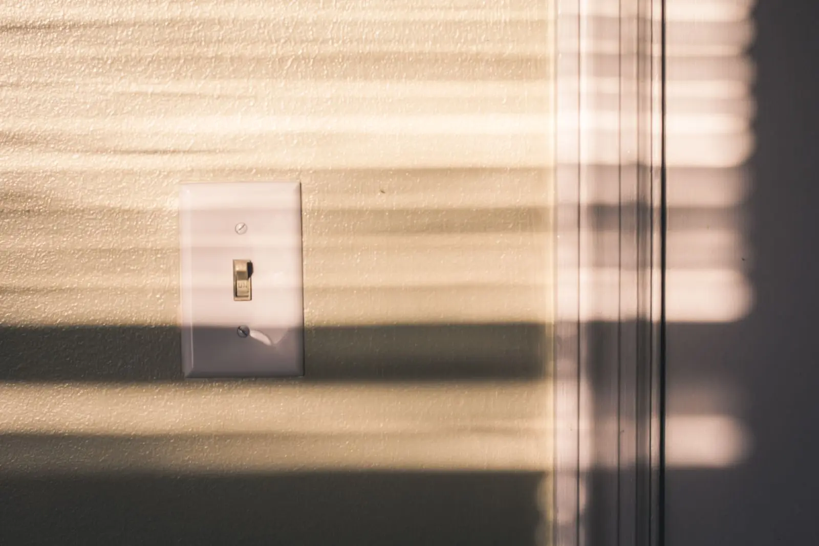 Toggle light switch on a wall striped with sunlight from window blinds