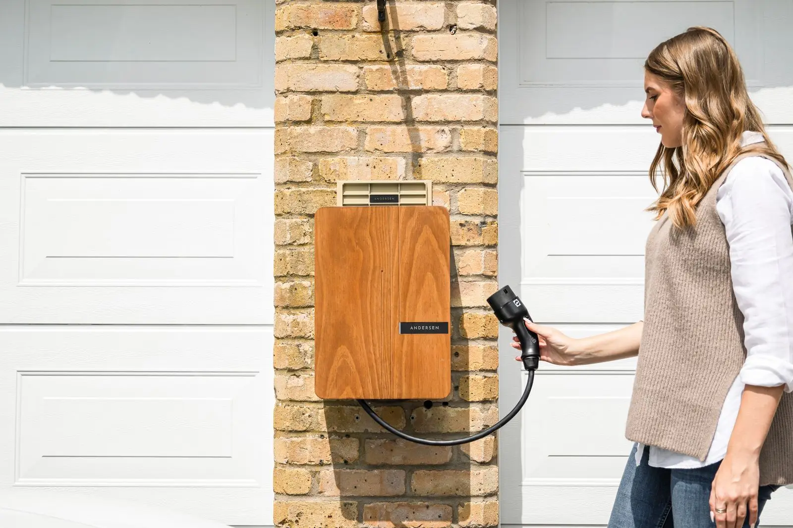 Woman taking the connector from a home EV charger mounted on a brick pillar beside a garage door