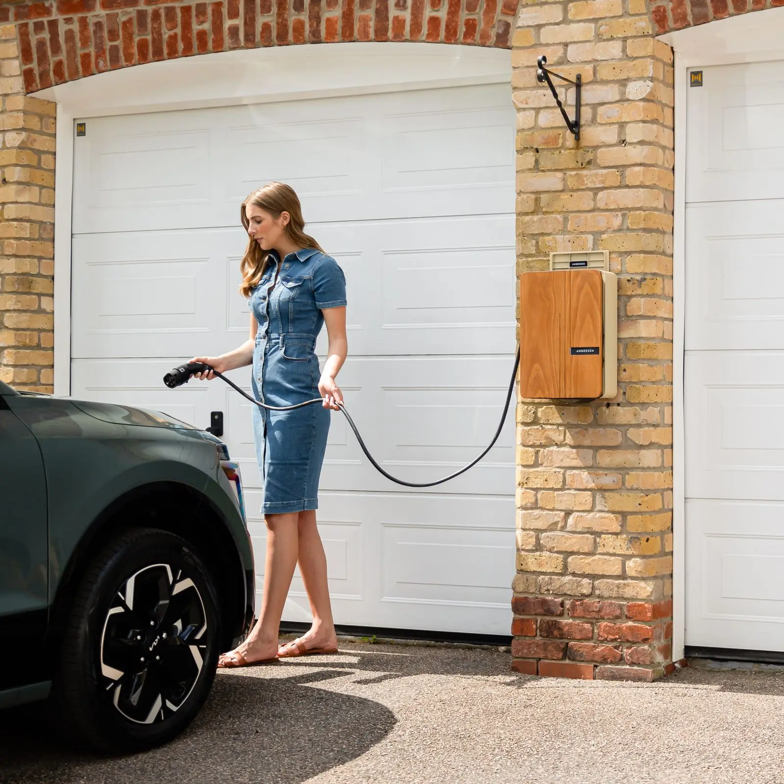 Woman carrying a charging cable from a wall-mounted home charger to a car in the driveway